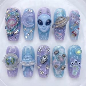 May include: A set of ten press-on nails featuring a space theme with glittery blue and purple gradients. Designs include a galaxy swirl, a crescent moon, a 3D alien head, a planet with rings, rockets, an astronaut helmet, a UFO, and asteroids.
