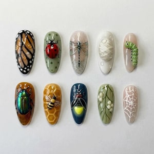 May include: A set of ten almond-shaped press-on nails featuring detailed 3D insect and nature designs. The collection includes a butterfly wing, a ladybug, a dragonfly, a white cocoon, a green caterpillar, a beetle, a bee on honeycomb, a firefly, a leafy branch, and a spider web.