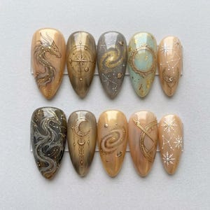 May include: A set of ten almond-shaped press-on nails featuring intricate celestial and fantasy designs. The artwork includes gold and white illustrations of dragons, galaxies, planets, moons, constellations, and snowflakes on beige, grey, and teal bases.
