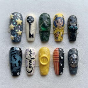 May include: A set of ten press-on nails featuring designs inspired by Coraline. The nails include illustrations of a black cat, a key, a yellow raincoat, stars, buttons, and the text Coraline, rendered in various colors including yellow, black, and blue.