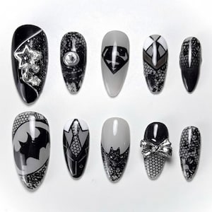 May include: A set of ten artificial nails in various designs. The nails feature black, gray, and white colors with embellishments. Designs include a star, a Superman logo, a Batman logo, and a bow.