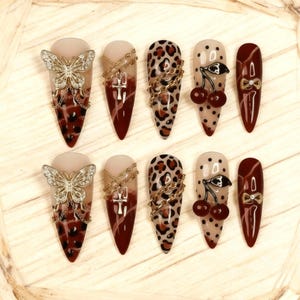 May include: A set of ten long, almond-shaped press-on nails. Designs include leopard print, gold chains, butterflies, cherries, and bows. The nails feature a mix of burgundy, nude, and gold accents.
