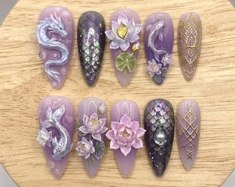 May include: A set of ten long, almond-shaped artificial nails. Each nail features intricate designs, including a dragon, koi fish, lotus flowers, and geometric patterns. The color palette includes shades of purple, silver, and gold, with a glossy finish.