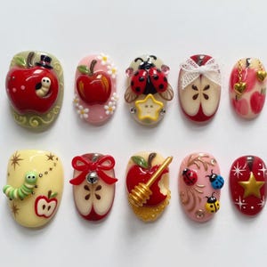 May include: A set of ten decorative nail art press-on pieces featuring various designs including red apples, ladybugs, a green caterpillar, a honey dipper, gold stars, white flowers, a white lace bow, a red bow with a bell, and gold heart charms.