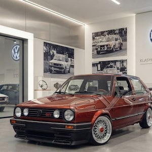 Könnte beinhalten: Ein metallisch kupferfarbener Volkswagen Golf GTI Schrägheck mit schwarzer Frontstoßstange, runden Scheinwerfern und silbernen Vielspeichenfelgen. Das Fahrzeug ist in einem Ausstellungsraum mit weißen Wänden und VW-Logo zu sehen.