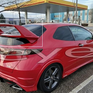 Peut inclure: Une voiture de sport rouge avec un grand aileron arrière. La voiture a des roues noires et des vitres teintées. Le véhicule est garé dans un parking près d'une station-service.