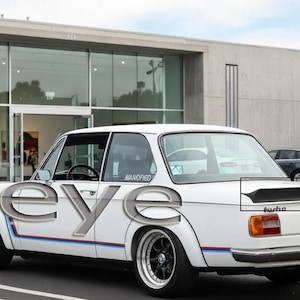 Puede incluir: Coche blanco vintage con ruedas negras y un alerón trasero. El coche tiene rayas azules, rojas y blancas a lo largo del lateral. La palabra "turbo" es visible en la parte trasera.