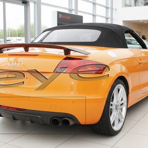 Op de afbeelding: Een oranje cabriolet met een zwarte spoiler en een zwart cabriodak. De auto heeft zilveren velgen en dubbele uitlaatpijpen. De auto staat in een showroom.