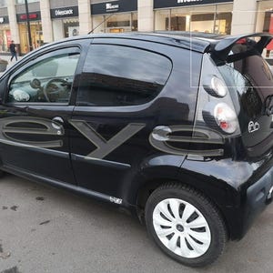Op de afbeelding: Een zwarte compacte auto met witte wieldoppen staat geparkeerd op straat. De auto heeft getinte ramen en een achterspoiler. Grote gestileerde letters zijn zichtbaar op de zijkant van de auto.