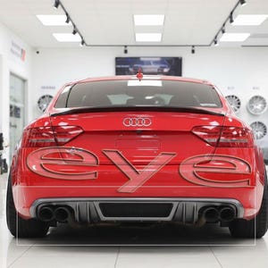 Pode incluir: Um carro desportivo Audi vermelho com a palavra "EYE" sobreposta na traseira. O carro tem um spoiler preto, tubos de escape duplos e um difusor traseiro preto. O fundo mostra um showroom com rodas.