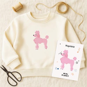 Puede incluir: Sudadera color crema con un gráfico de caniche rosa. Una transferencia de planchado a juego con el texto "Bügelbild" y "MINI PLANET" se muestra junto a la sudadera. La imagen también incluye tijeras, hilo y cuerda.