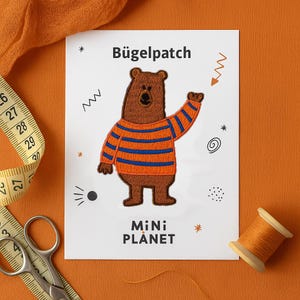 Könnte beinhalten: Ein brauner Bärenaufnäher mit blau-orange gestreiftem Pullover. Der Aufnäher hat den Text "Bügelpatch" oben und "MiNi PLANET" unten. Auf weißem Hintergrund mit orangefarbenen Akzenten.