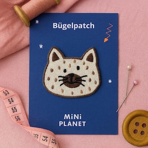 May include: A cat-shaped iron-on patch with a white base and brown spots, ears, and details. The patch is on a blue card with the text "Bügelpatch" and "MiNi PLANET". A pink measuring tape, pins, and a wooden button are also visible.