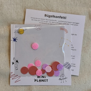 Könnte beinhalten: Eine transparente Plastikverpackung mit bunten, glitzernden Konfetti in Rosa-, Lila- und Brauntönen. Die Verpackung hat ein Weltraum-Design mit einer Rakete und den Worten "MINI PLANET". Eine weiße Karte mit deutschem Text befindet sich hinter der Verpackung.