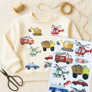 May include: Cream-colored sweatshirt with colorful watercolor illustrations of various vehicles, including a fire truck, helicopter, and police car. A sheet of matching iron-on transfers is next to the sweatshirt. A pair of scissors and thread are also in the image.