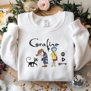 May include: White sweatshirt featuring the word "Coraline" in black script, along with cartoon illustrations of characters from the movie. The design includes a black cat, a character in a blue coat, and other related imagery.