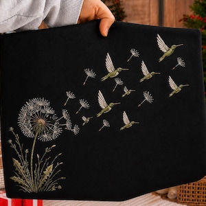 Könnte beinhalten: Ein schwarzer Stoffartikel mit einer Stickerei, die eine Pusteblume zeigt, deren weiße Samen davonwehen und sich in mehrere kleine grüne Kolibris im Flug verwandeln. Die Stickerei verwendet Fäden in Grün-, Weiß- und Brauntönen.