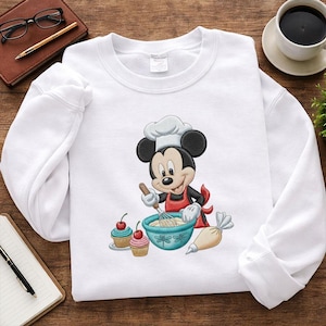 Peut inclure: Sweat-shirt blanc avec une image brodée de Mickey Mouse en tant que chef, portant un chapeau de chef et un tablier rouge, fouettant dans un bol bleu. Deux cupcakes et une poche à douille sont également brodés. Un design amusant et fantaisiste.