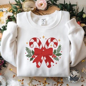 May include: White sweatshirt featuring a festive candy cane design. Two red and white striped candy canes are crossed with a red bow, surrounded by green leaves and red berries. Gold stars add a touch of sparkle to this holiday-themed apparel.