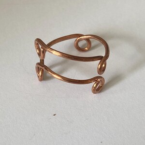 May include: A handcrafted copper wire ring with a unique, open design. The ring features a series of curved lines and swirls, creating an artistic and elegant look. The copper has a warm, reddish-brown hue, and the ring is displayed on a white surface.