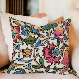 Puede incluir: Un cojín decorativo con un colorido estampado floral. El cojín presenta un fondo color crema con un diseño repetido de flores y hojas rosas, azules, amarillas y verdes. El cojín está colocado sobre un sillón rosa claro.