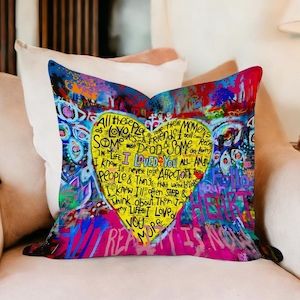 Könnte beinhalten: Ein dekoratives herzförmiges Kissen mit einem lebendigen Graffiti-Design. Das Kissen hat ein gelbes Herz in der Mitte mit handgeschriebenem Text, umgeben von bunten Farbspritzern und Wörtern in verschiedenen Farben. Das Kissen liegt auf einem beigen Sofa.