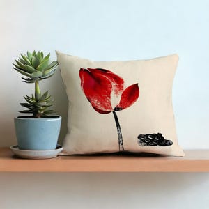 Peut inclure: Coussin carré crème avec un grand motif de tulipe rouge. La tulipe a une tige noire et une forme noire abstraite à la base. Une plante succulente en pot est à gauche.