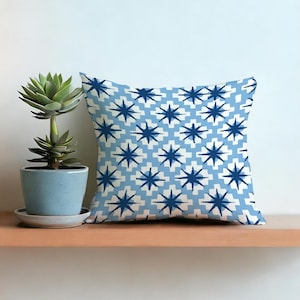 Op de afbeelding: Vierkant sierkussen met een blauw en wit sterrenpatroon. Het kussen ligt op een houten plank naast een vetplant in een blauwe pot.