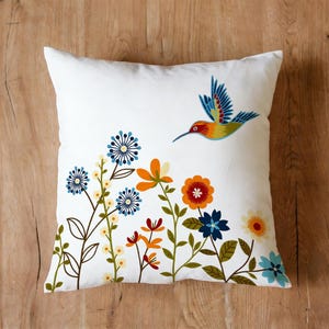 May include: White square throw pillow with a colorful floral and hummingbird design. The flowers are in shades of blue, orange, and red, with green stems and leaves. A hummingbird with blue, yellow, and red feathers is flying above the flowers.