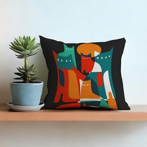 Puede incluir: Cojín cuadrado negro con un diseño abstracto de gatos en naranja, turquesa y rojo con detalles blancos. A la izquierda, una planta suculenta en una maceta azul.