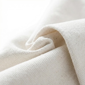 May include: Close-up of a textured, off-white fabric with visible weave. The fabric is folded and curled, creating soft shadows and highlighting the material's texture. The image emphasizes the natural, organic quality of the textile.