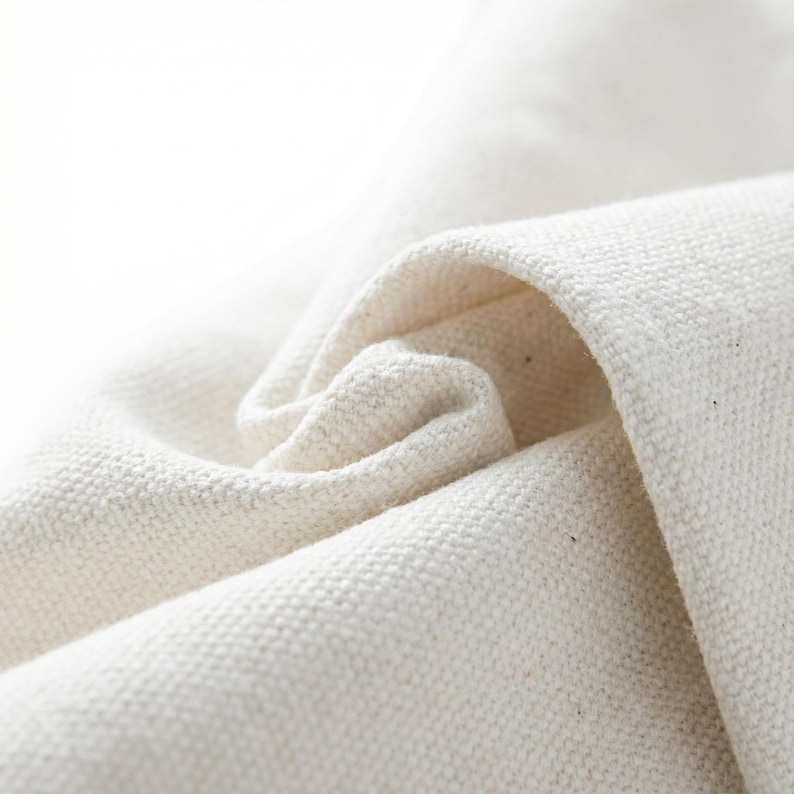 May include: Close-up of a textured, off-white fabric with visible weave. The fabric is folded and curled, creating soft shadows and highlighting the material's texture. The image emphasizes the natural, organic quality of the textile.