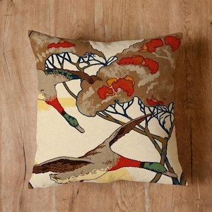May include: Square decorative pillow with a nature-inspired design. The pillow features a cream-colored background with a print of flying ducks, trees, and foliage in shades of brown, red, and blue. The pillow is placed on a wooden surface.
