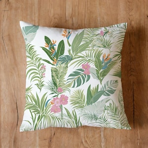 Può includere: Cuscino quadrato bianco con motivo floreale tropicale. Il design presenta foglie verdi, fiori rosa e fiori di uccelli del paradiso arancioni e blu. Il cuscino è posto su una superficie di legno.