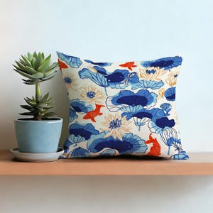 以下が含まれることがあります： クリーム色の背景に、青い蓮の葉、オレンジ色の鯉、白い花が描かれた正方形の装飾用クッション。 青い鉢に入った多肉植物が木製の棚に置かれています。