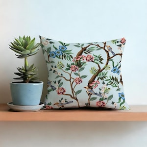 Op de afbeelding: Vierkant sierkussen met een lichtblauwe achtergrond met een bloemen- en vogelpatroon. Het ontwerp bevat roze en blauwe bloemen, groene bladeren en bruine takken. Een vetplant in een pot staat links op een houten plank.