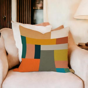 May include: A decorative pillow with a geometric design. The pillow features a patchwork of colors including orange, teal, mustard yellow, and gray. The pillow is rectangular and rests on a white armchair.