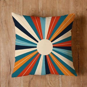 May include: Square throw pillow with a retro sunburst design. The pillow features a cream-colored center circle with radiating stripes in teal, navy, orange, red, and cream. The pillow is placed on a wooden surface.
