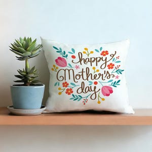 Op de afbeelding: Wit vierkant kussen met de tekst "happy Mother's day" in bruin schrift, omringd door kleurrijke bloemen en bladeren. Een vetplant in pot staat links op een houten plank.