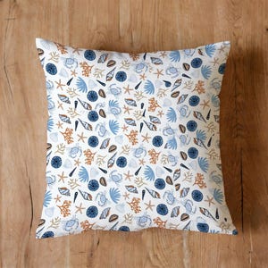 Puede incluir: Cojín cuadrado con un estampado de temática marina sobre fondo blanco. El diseño incluye cangrejos azules, conchas marinas, estrellas de mar y coral en tonos azules, marrones y blancos. El cojín está sobre una superficie de madera.