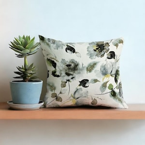 Puede incluir: Cojín decorativo cuadrado con un estampado floral en tonos grises, negros y verdes sobre fondo blanco. Se encuentra sobre una estantería de madera, junto a una pequeña suculenta en maceta azul claro. Medidas: aprox. 40 cm x 40 cm.