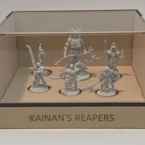 Pode incluir: Uma caixa de exibição de madeira e acrílico transparente com seis figuras em miniatura cinzentas. As figuras representam guerreiros com várias armas. O texto "Kainan's Reapers" está gravado na frente da caixa.