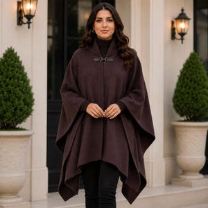 May include: A dark brown hooded cape featuring a structured collar with a black metal toggle clasp closure. The garment has long, wide sleeves with fitted cuffs, a draped silhouette, and a smooth, solid-coloured knit texture throughout the piece.
