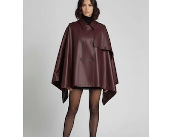 May include: A burgundy leather cape with a double-breasted front and a classic collar. The cape features a button closure and a flowing silhouette. The model is wearing a black turtleneck and black tights. The cape is a stylish outerwear piece.