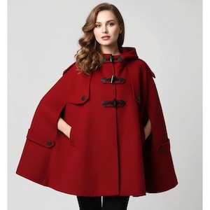 May include: A red hooded cape with black toggle closures and pockets. The cape is made of a thick material and has a classic design. The cape is a stylish outerwear option for cooler weather.