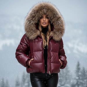K&ouml;nnte beinhalten: Eine bordeauxfarbene Steppjacke mit Kapuze mit Pelzbesatz. Die Jacke hat ein gl&auml;nzendes Finish und einen Rei&szlig;verschluss. Die Kapuze ist hellbraun. Der Hintergrund ist eine verschneite Szene im Freien.