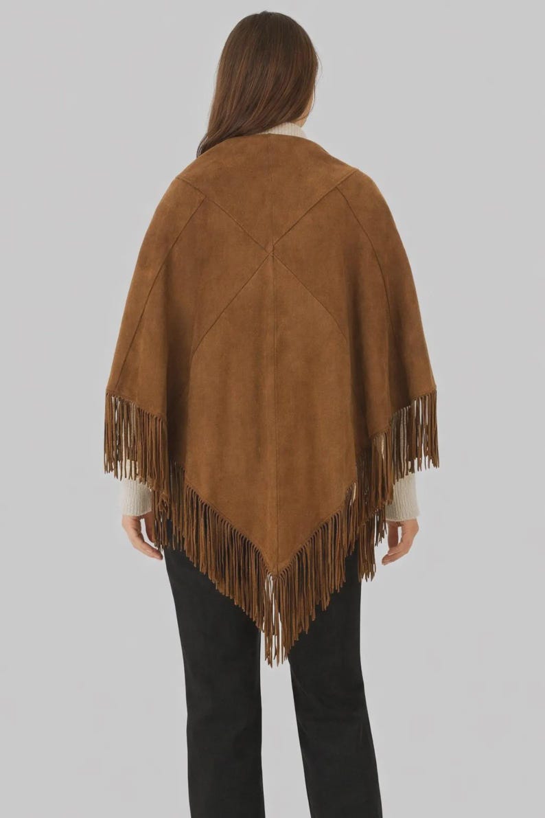 Op de afbeelding: Bruine su&egrave;de poncho met franjes. De poncho heeft een vierkante vorm met een gestikt ontwerp op de rug. De franjes hangen langs de onderkant van de poncho.