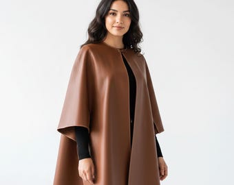 May include: A brown leather cape with a round neckline and a front opening. The cape has a flowing design and is worn over a black top. The background is plain white.