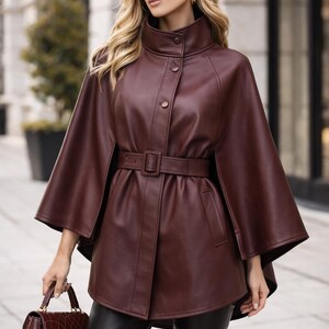 May include: A burgundy leather cape coat with a high collar and a matching belt. The coat has a button closure and flared sleeves. A matching burgundy handbag is held in the hand.