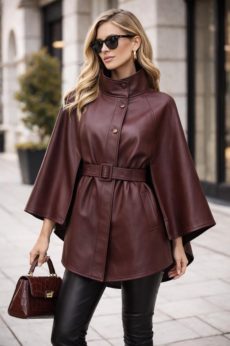 May include: A burgundy leather cape coat with a high collar and a matching belt. The coat has a button closure and flared sleeves. A matching burgundy handbag is held in the hand.